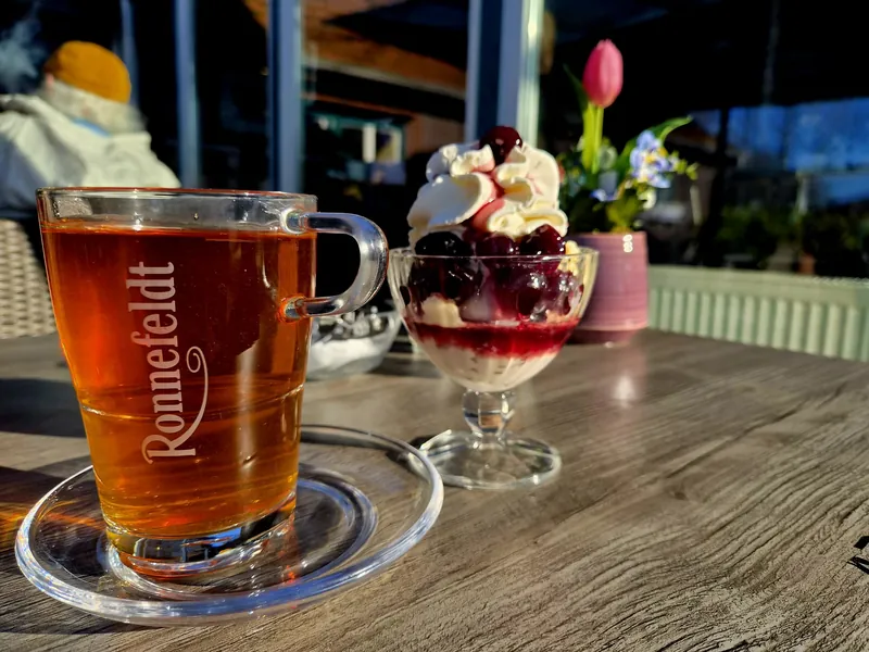 Ein Glas Schwarztee und ein Kirschbecher mit Sahne auf einem Holztisch in der Abendsonne